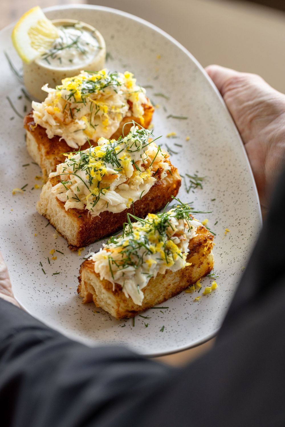 Hand holding a plate of toasted bread topped with crab meat, lemon zest, and fresh dill, served with a dipping sauce.