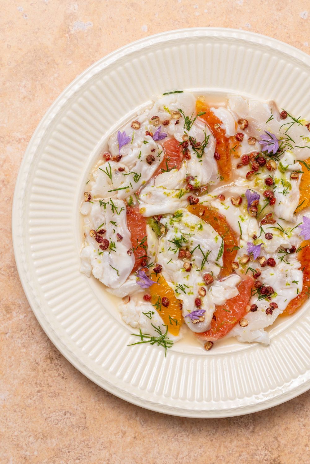 Elegant white plate with citrus salad featuring grapefruit, orange segments, cream dressing, dill, edible flowers, and pink peppercorns.