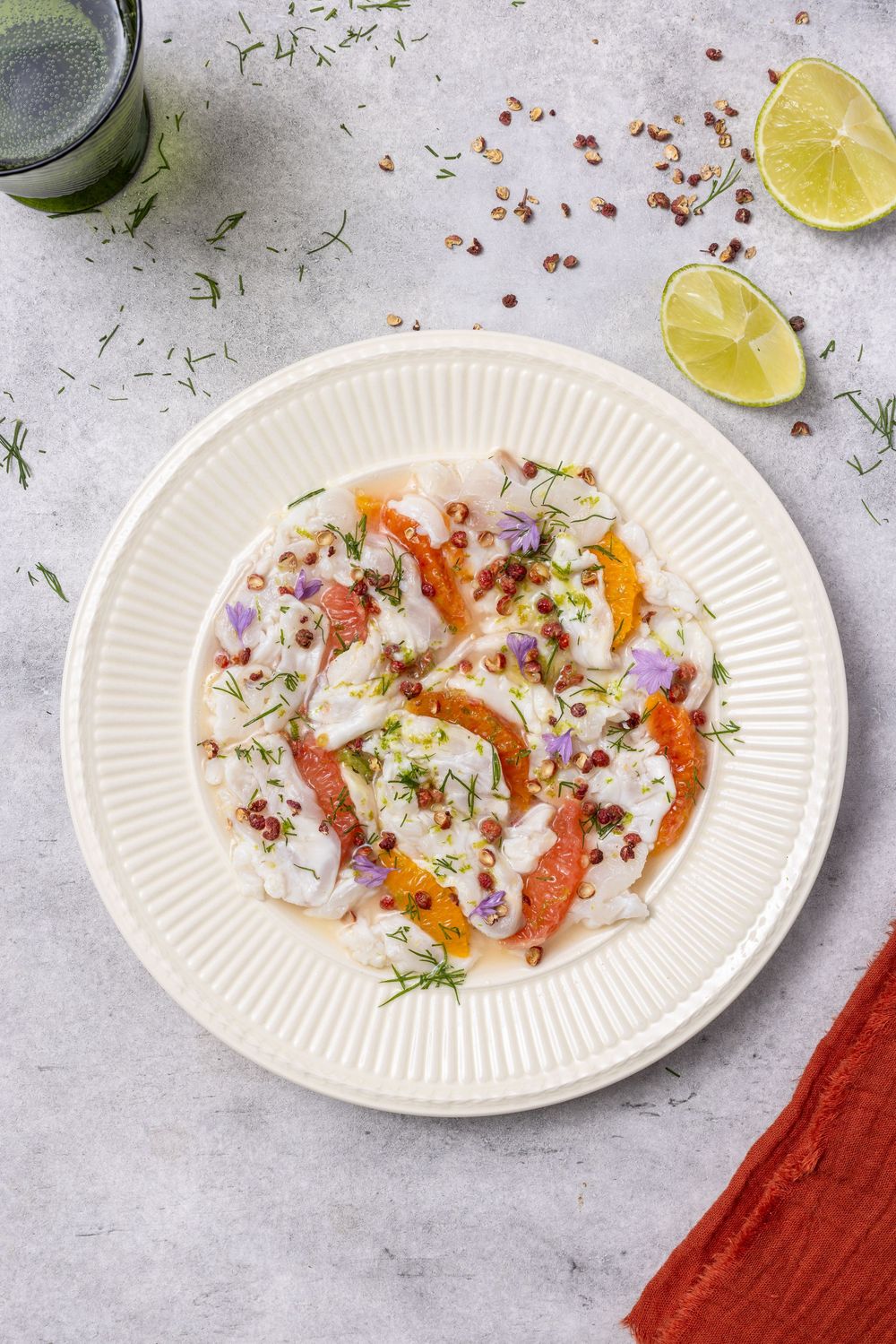 Elegant fish carpaccio with citrus segments, fresh dill, pink peppercorns, and edible flowers on a white plate.