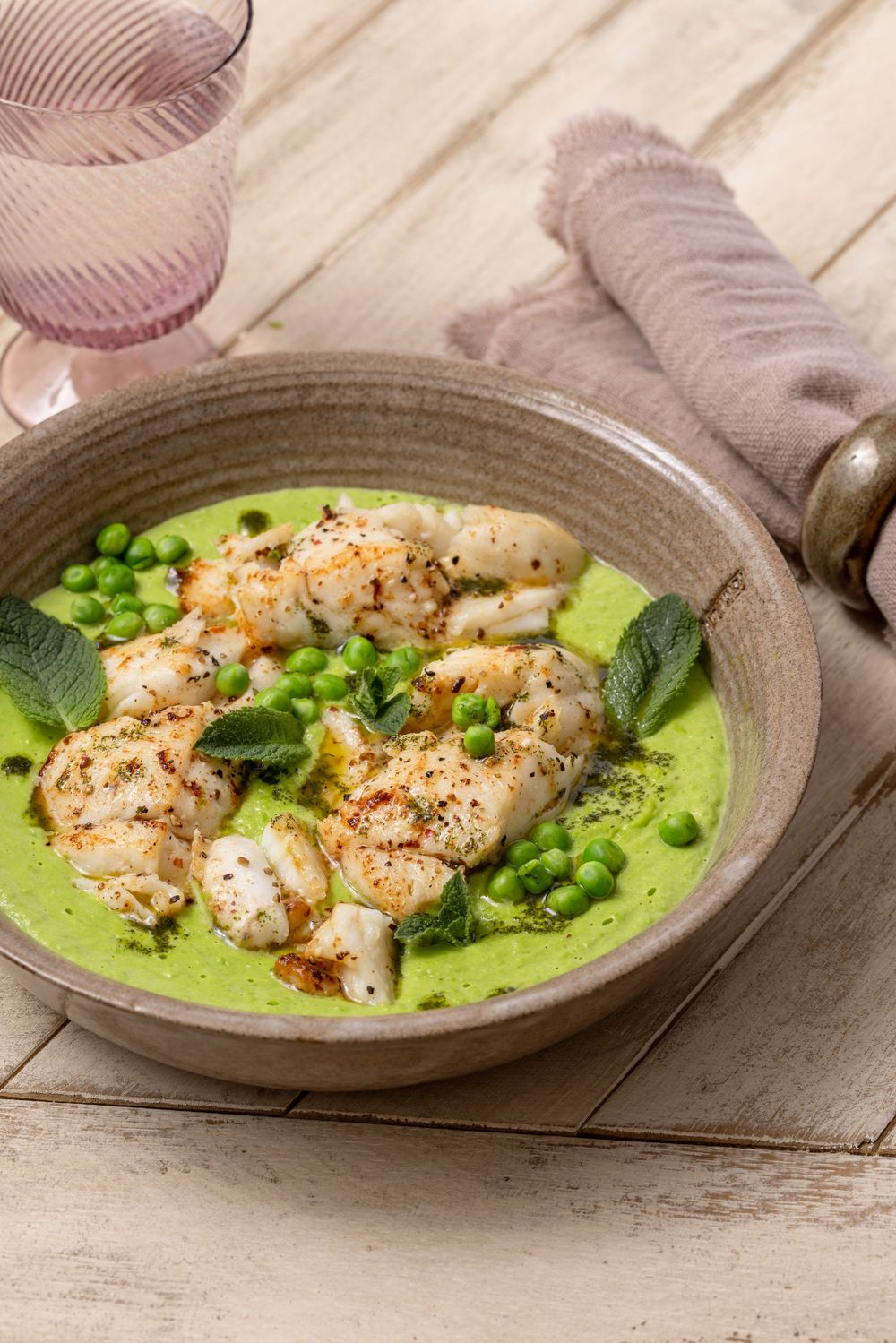 Seared cod fillets served in a vibrant green pea cream sauce with fresh peas and sage leaves in a rustic ceramic bowl.