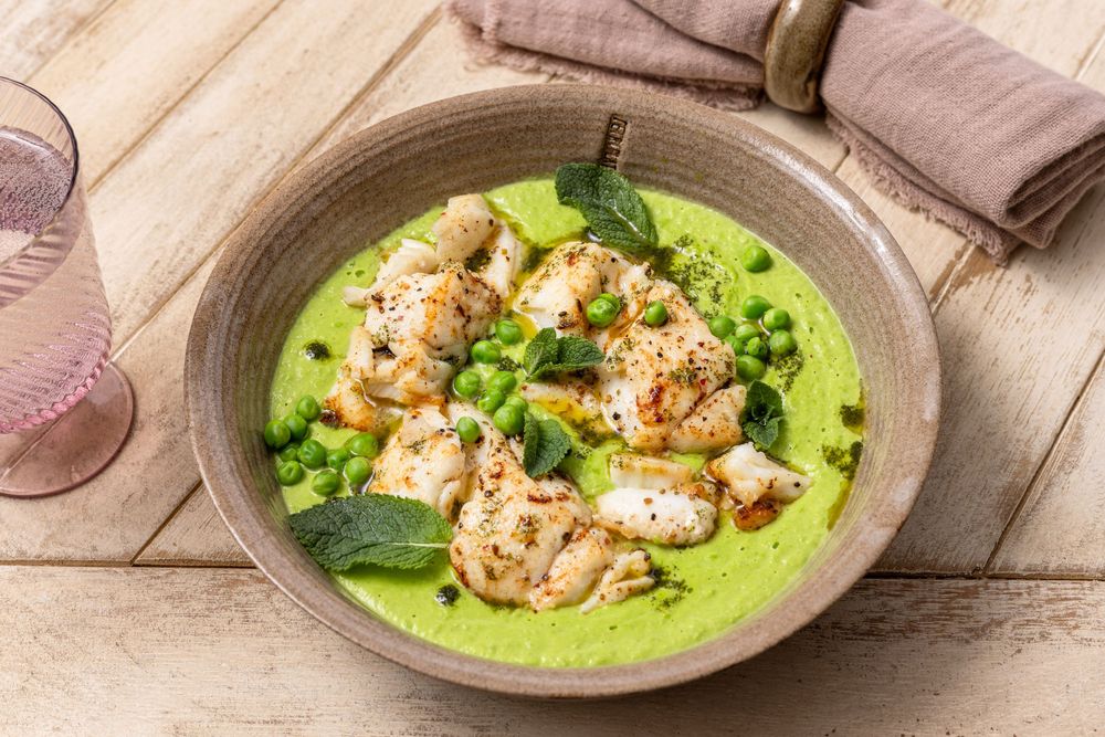 Grilled chicken pieces served in a vibrant green pea puree soup, garnished with fresh herbs in a rustic ceramic bowl.