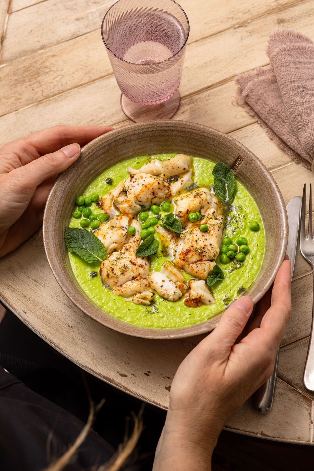 Hands holding a bowl of vibrant green pea soup topped with seared scallops, peas, and fresh herbs.