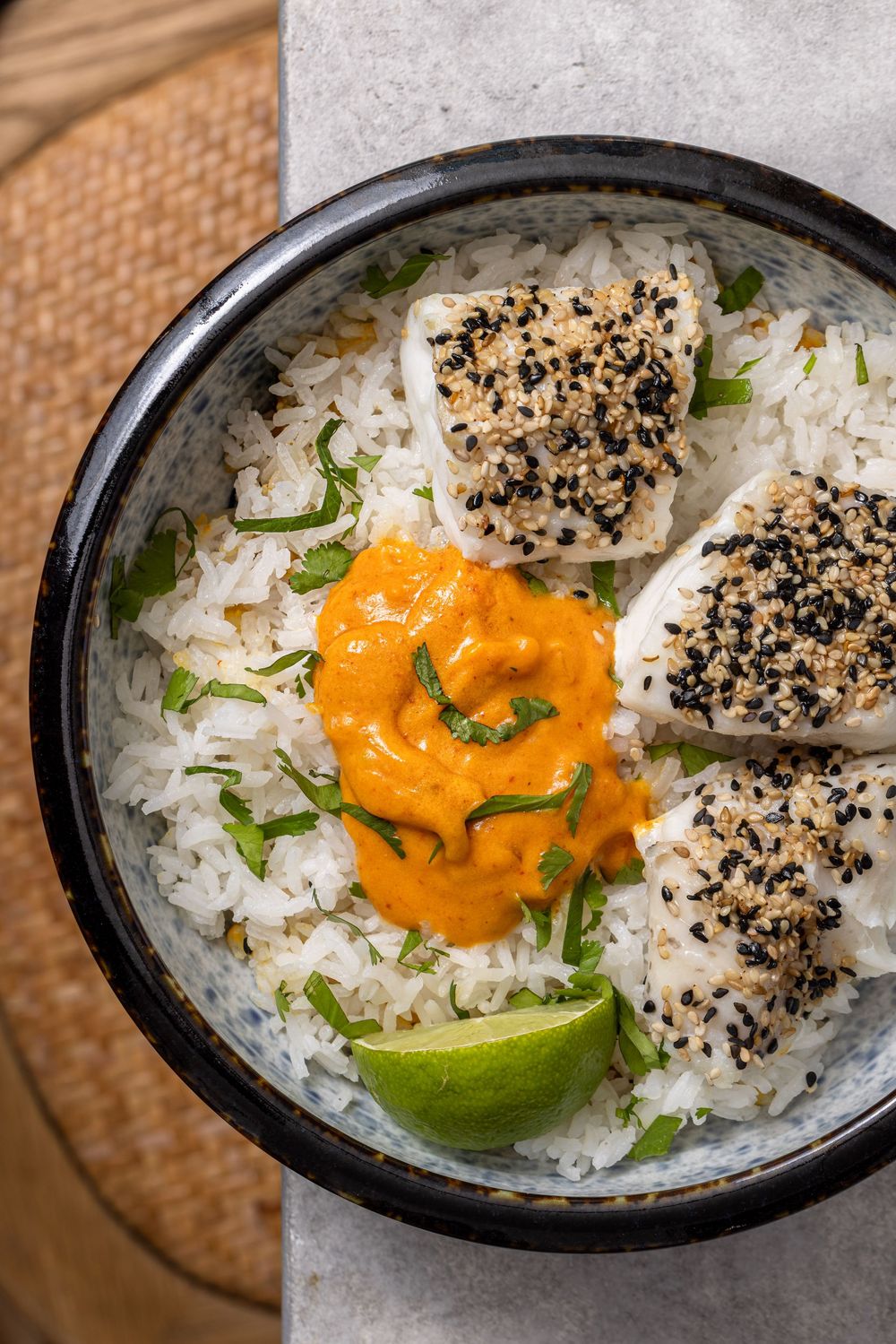 Sesame-crusted fish fillets served over white rice with orange sauce, fresh herbs, and lime wedge in a ceramic bowl.