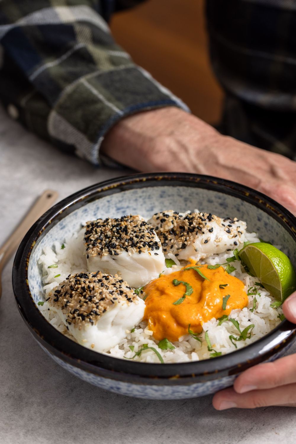 Sesame-crusted cod fillet served over rice with orange sauce and lime wedges in a ceramic bowl held by hands.