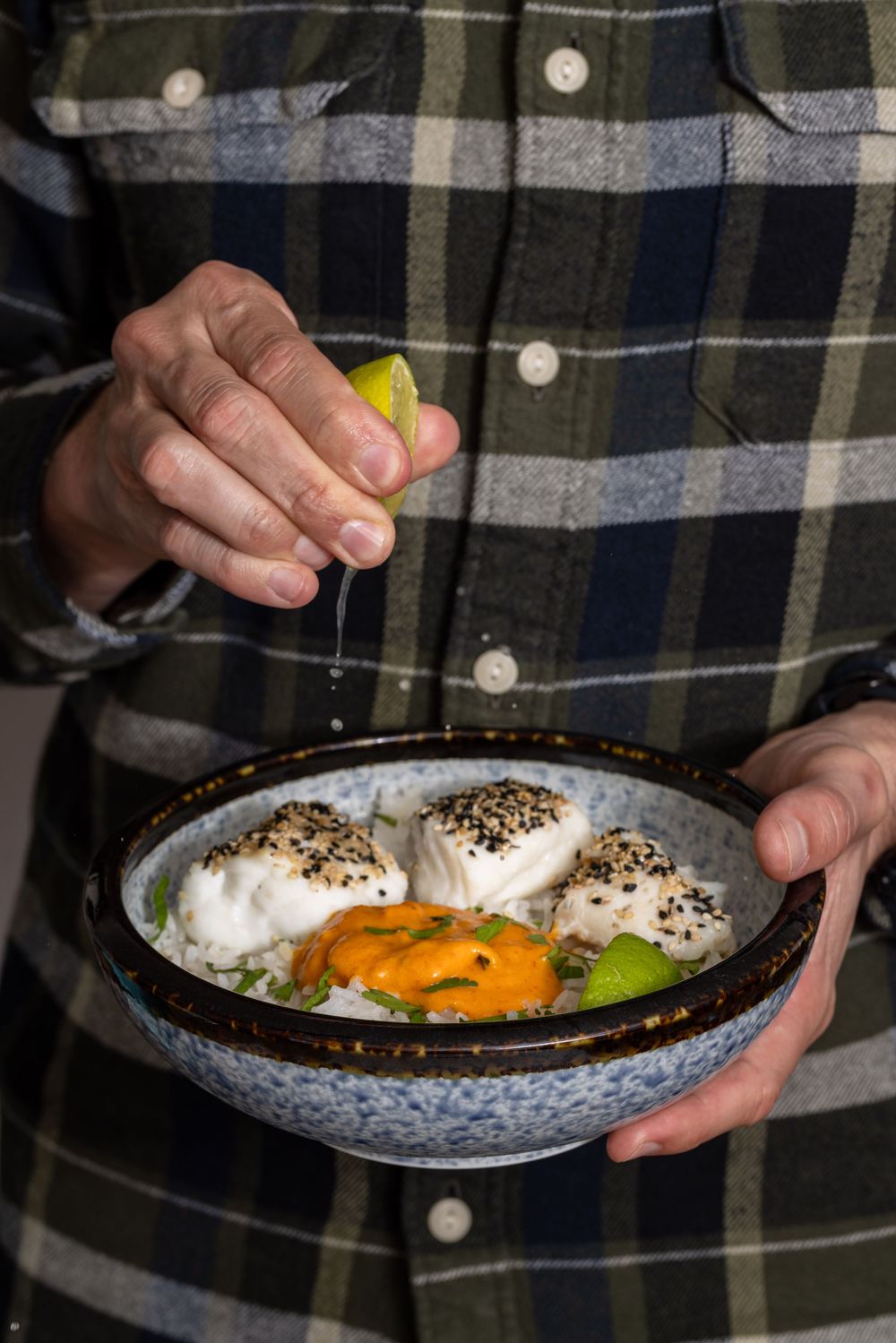Person squeezing lime over a bowl of sesame-crusted fish with rice, avocado, and orange sauce.