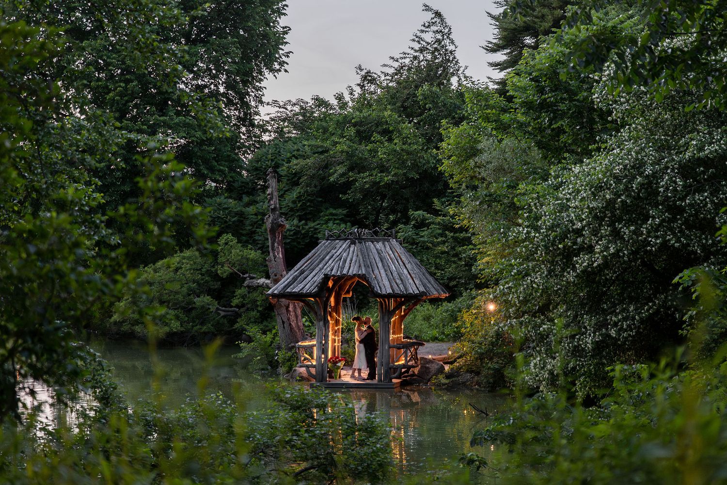 A cozy illuminated gazebo nestled among lush green trees at dusk.
