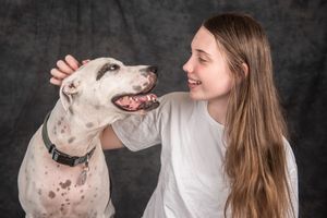 A smiling teenage girl with long brown hair pets a white Great Dane dog against a dark gray background.