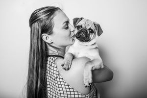 Black and white photo of a young woman kissing her pug puppy against a white background.