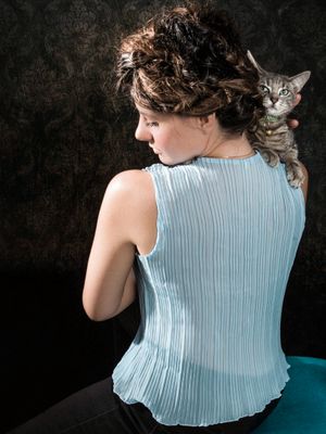 Woman in light blue sleeveless top seen from behind, holding a tabby cat on her shoulder against dark background.