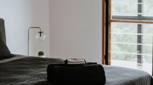 Minimalist bedroom with dark bedding, folded towels, brass lamp, and large window overlooking lush greenery.