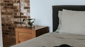 Modern bedroom with gray upholstered bed, linen bedding, wooden nightstand, geometric terrarium, and exposed brick wall.