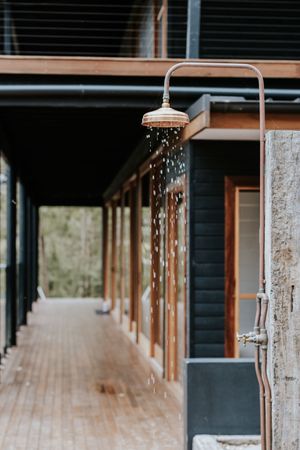 Outdoor rain shower head with water flowing, mounted on rustic wooden deck beside dark modern building exterior.