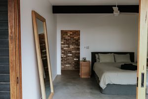 Modern bedroom with concrete floors, brick accent wall, ceiling fan, large mirror, and neutral linen bedding.