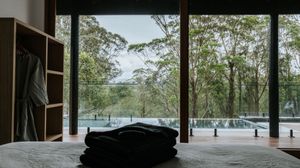 Modern bedroom with floor-to-ceiling windows overlooking lush forest and pool, featuring folded dark linens on bed.