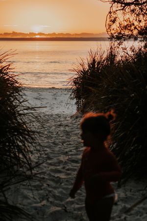 Child silhouetted against a golden beach sunset, framed by coastal grasses with calm ocean waters glowing in the background.