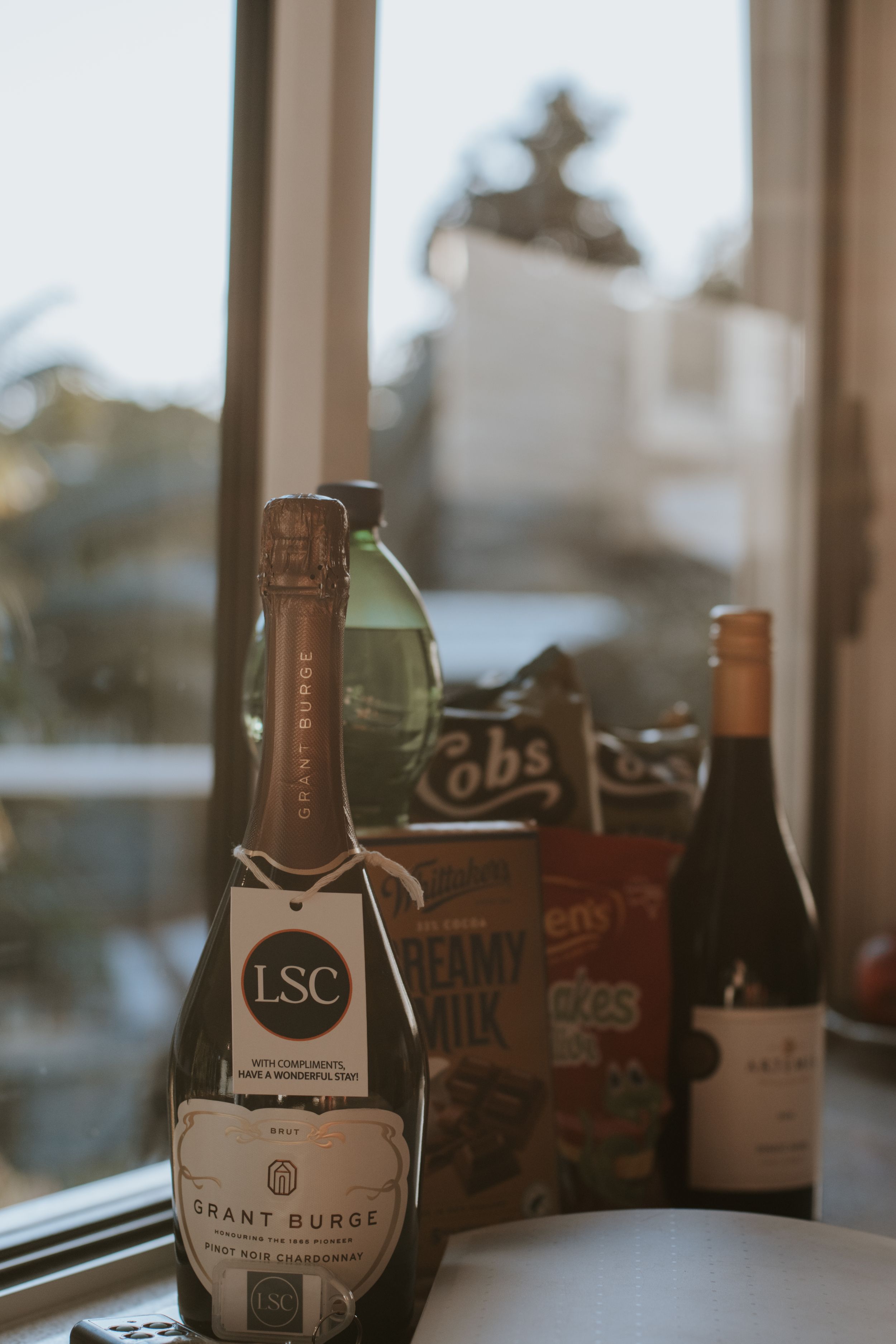 Grant Burge sparkling wine bottle with LSC tag displayed beside Cadbury Creamy Milk chocolate and snacks on a windowsill.