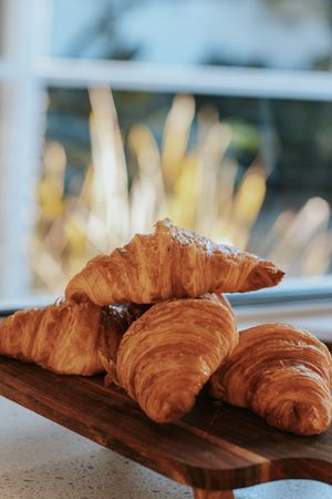 Golden flaky croissants stacked on a rustic wooden board by a sunlit window, perfect for a bakery breakfast spread.