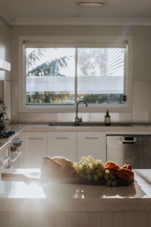 Sunlit white kitchen with fruit bowl on marble island, featuring green grapes and bread near sink with window view.