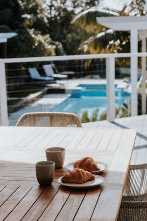Croissants and coffee cups on a wooden table overlooking a serene outdoor pool and lush garden.