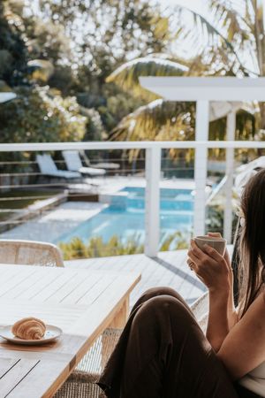 Woman relaxing on a sunny poolside terrace with a croissant and coffee, surrounded by lush tropical greenery.
