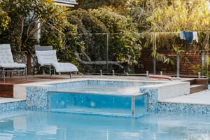 Luxury outdoor pool with glass-walled spa, lounge chairs, and lush greenery in a modern backyard setting.