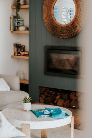 Cozy living room with green fireplace, stacked logs, round rattan mirror, built-in shelves, and a small white table with succulent.