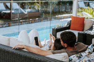 Couple relaxing on outdoor patio sofa by pool, using smartphones on a sunny day.