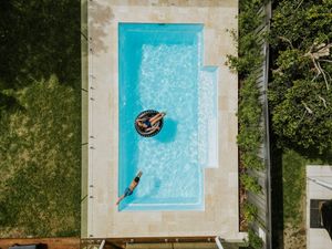 Aerial view of a rectangular backyard swimming pool with two people relaxing, one on a float, surrounded by green grass.
