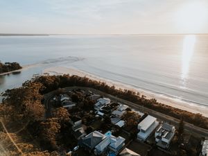 Aerial drone view of a coastal beach town with houses surrounded by trees and golden sunlight reflecting on calm ocean waters.