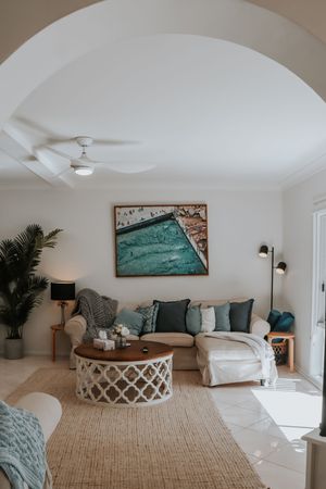 Bright coastal living room with beige sectional sofa, round white coffee table, teal artwork, and ceiling fan under arched ceiling.