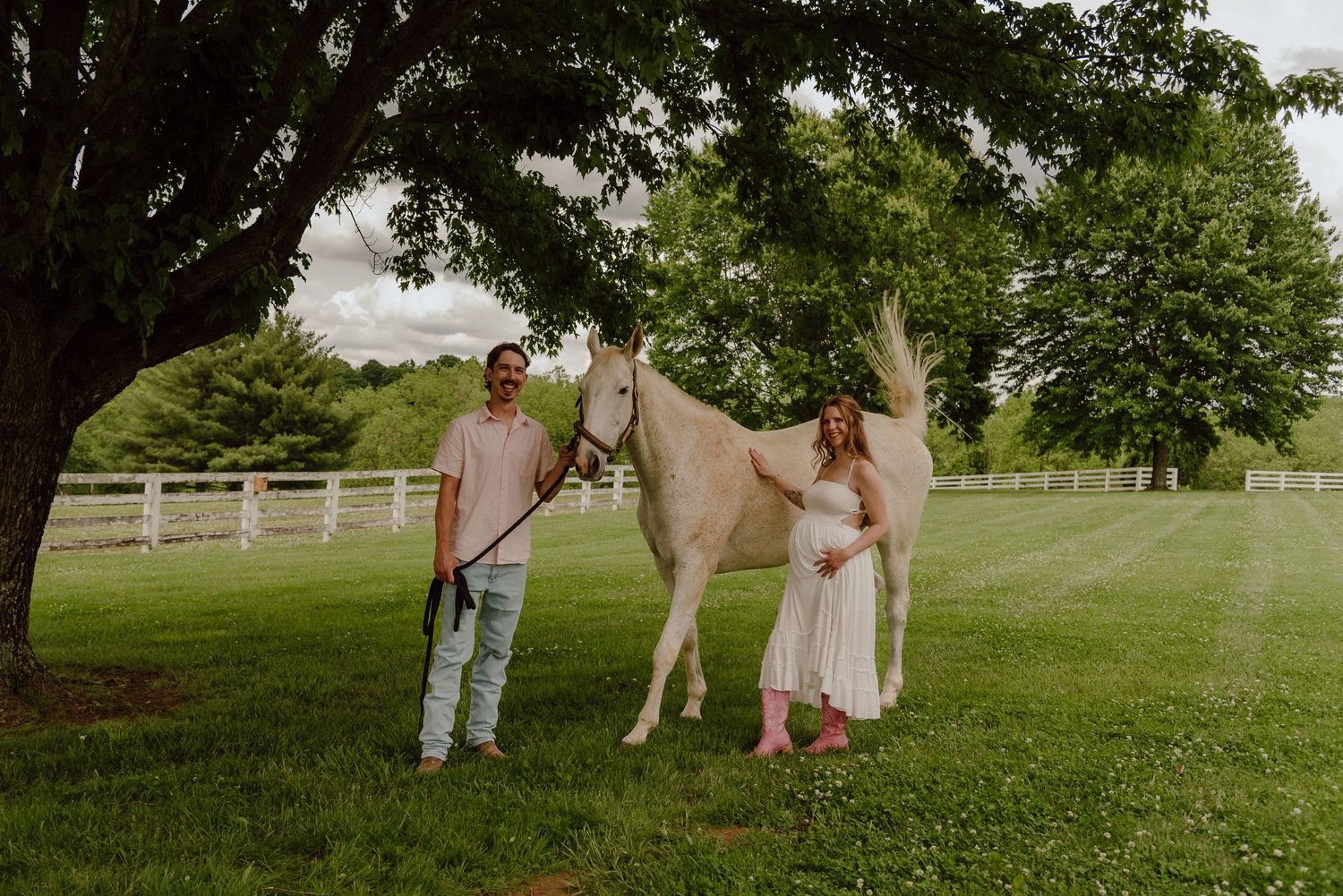 A couple stands with a white horse on a lush green farm under a large tree near a white fence.