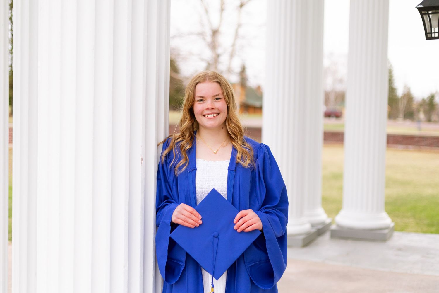 Young woman in blue graduation gown and cap smiling between white columns outdoors.
