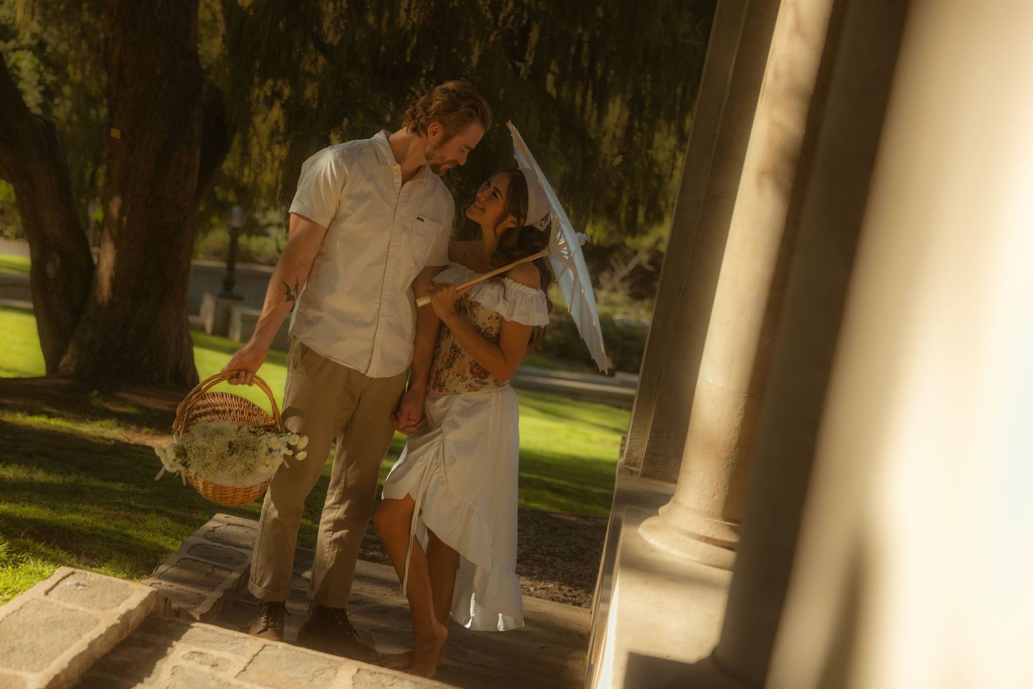 Couple in wedding attire sharing a romantic moment outdoors, bride in white dress with parasol, groom in casual dress pants.