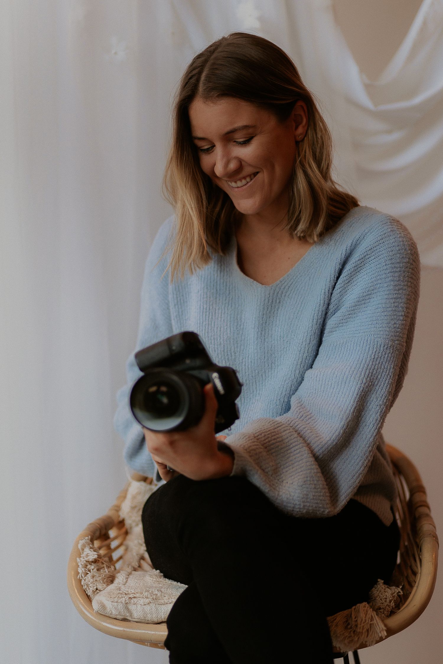 Smiling woman in blue knit sweater sitting in rattan chair, holding a DSLR camera in a bright studio.