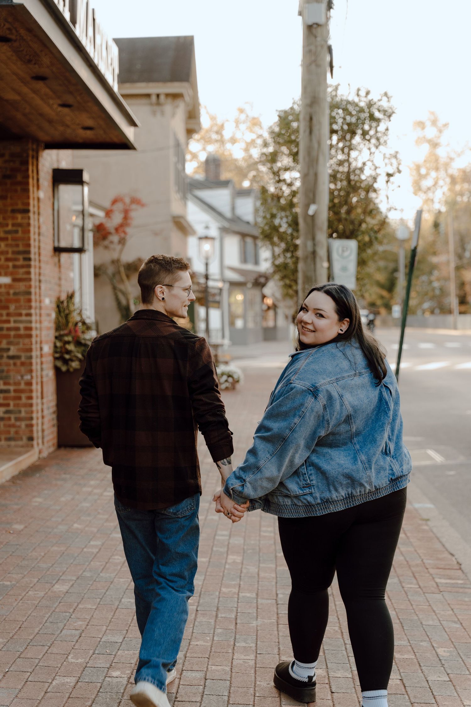 A couple holds hands while walking on a brick sidewalk, the woman in a denim jacket smiling back at the camera.
