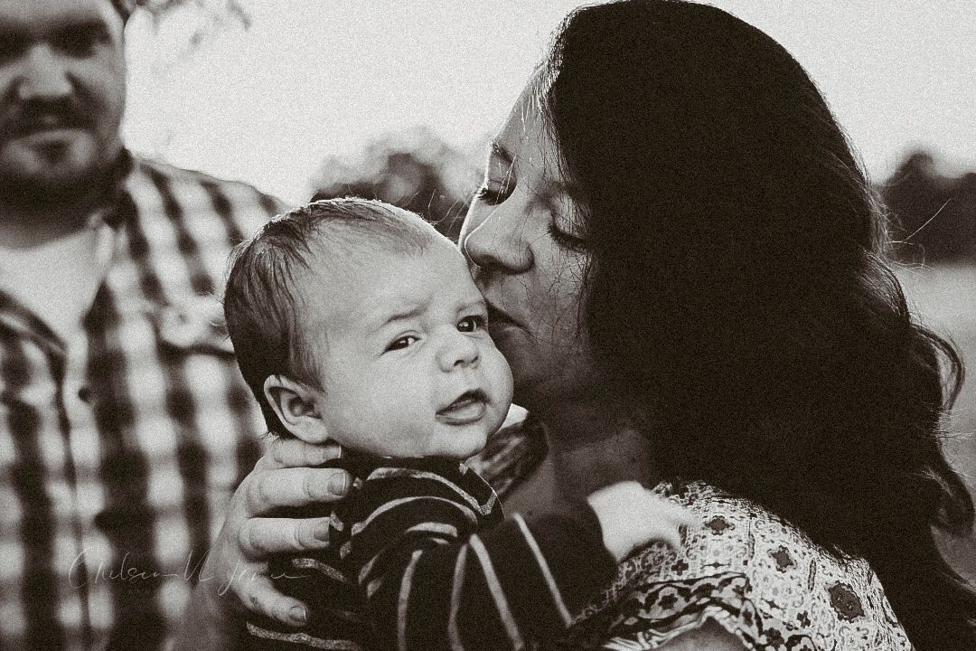 Black and white photo of a mother kissing her baby while a man stands beside them outdoors.