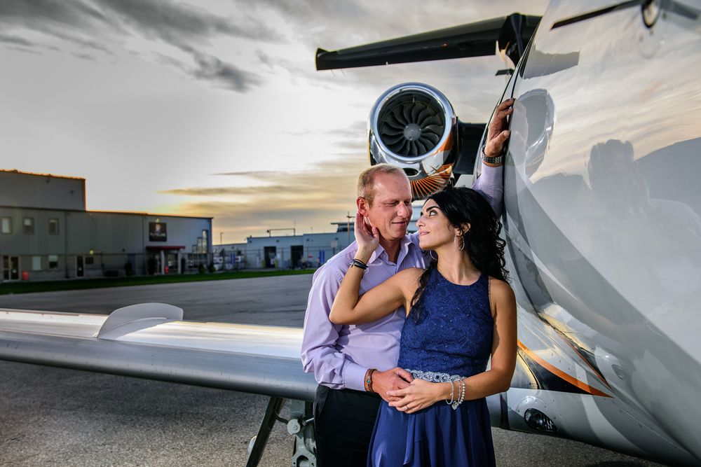 Couple embracing beside a private jet engine at sunset on an airport tarmac, woman in blue dress.