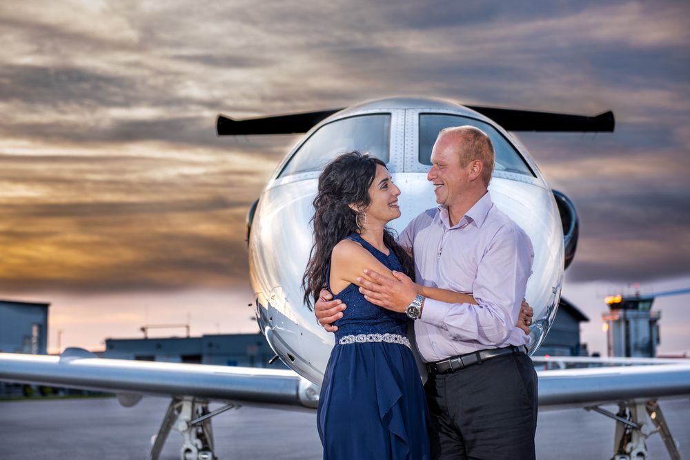 Couple embracing and smiling in front of a private jet nose at golden sunset on airport tarmac.