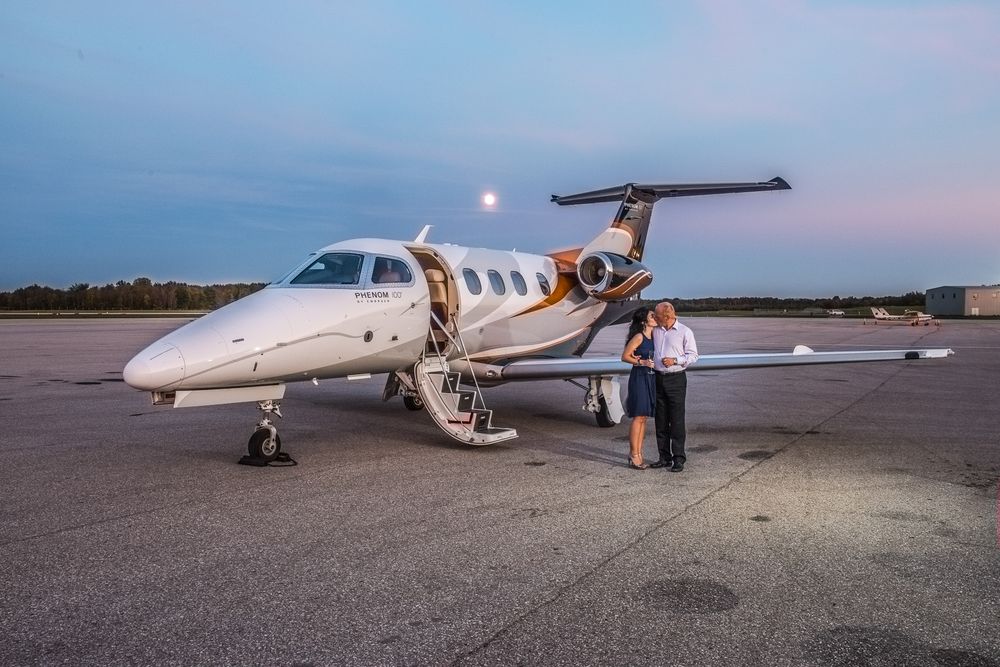 Couple standing together beside a white private jet on a tarmac at dusk.