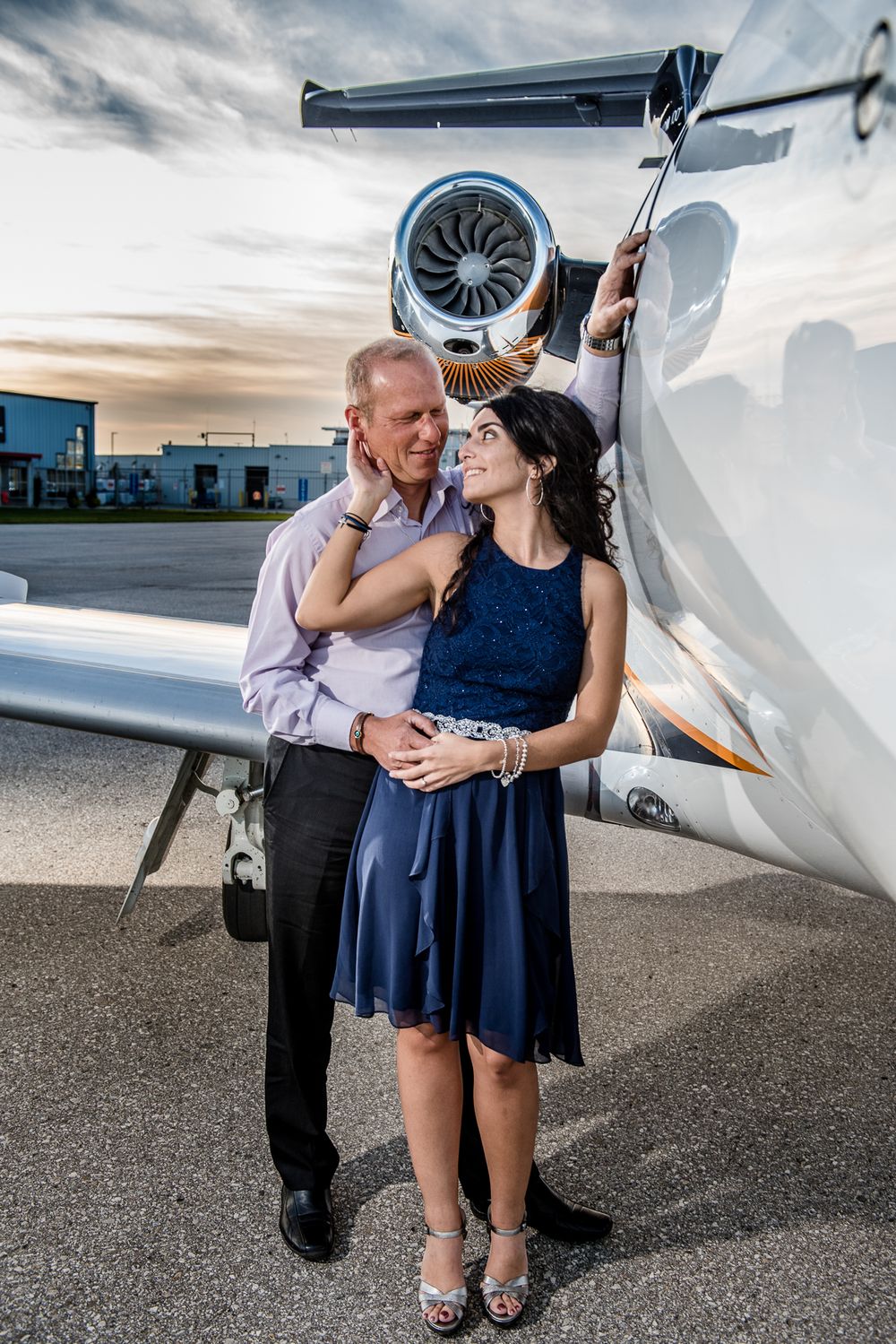 Elegant couple posing next to a white private jet at dusk, woman in blue dress and heels smiling.