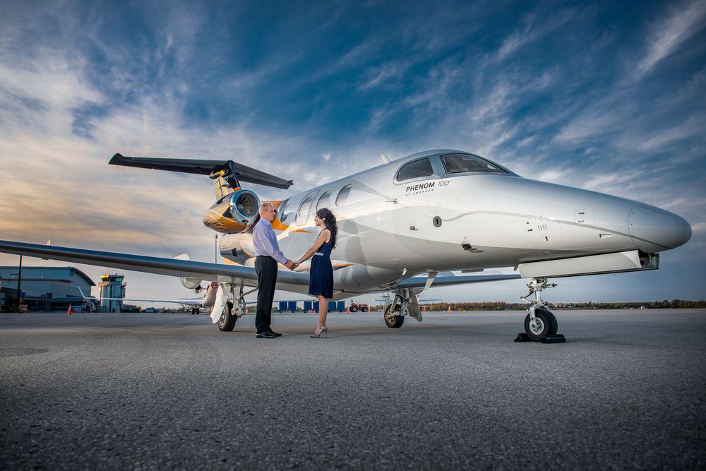 Couple standing on airport tarmac beside a sleek white private jet under a dramatic sunset sky.