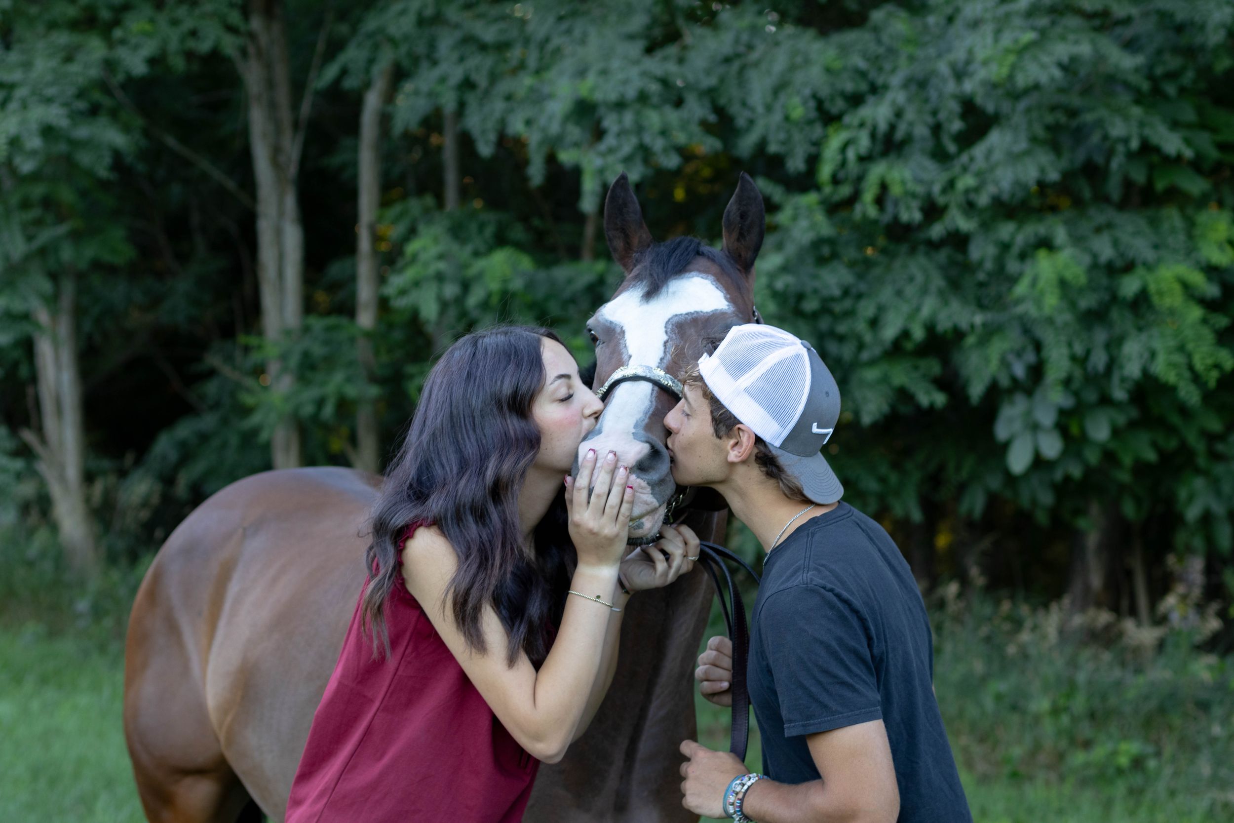 A couple kisses a brown horse on the nose outdoors, surrounded by lush green trees.