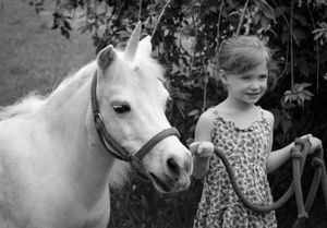 A young smiling girl in a floral dress stands beside a white pony in a black and white photo.