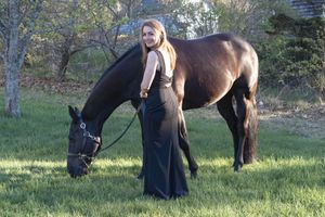 Woman in black gown poses with bay horse grazing on green grass outdoors.
