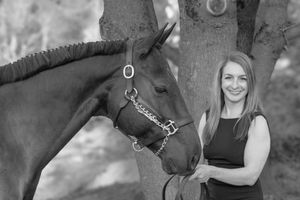 Black and white photo of smiling woman standing beside bay horse near tree.