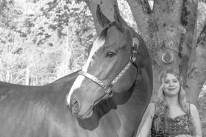 Black and white portrait of blonde woman posing with bay horse by tree.