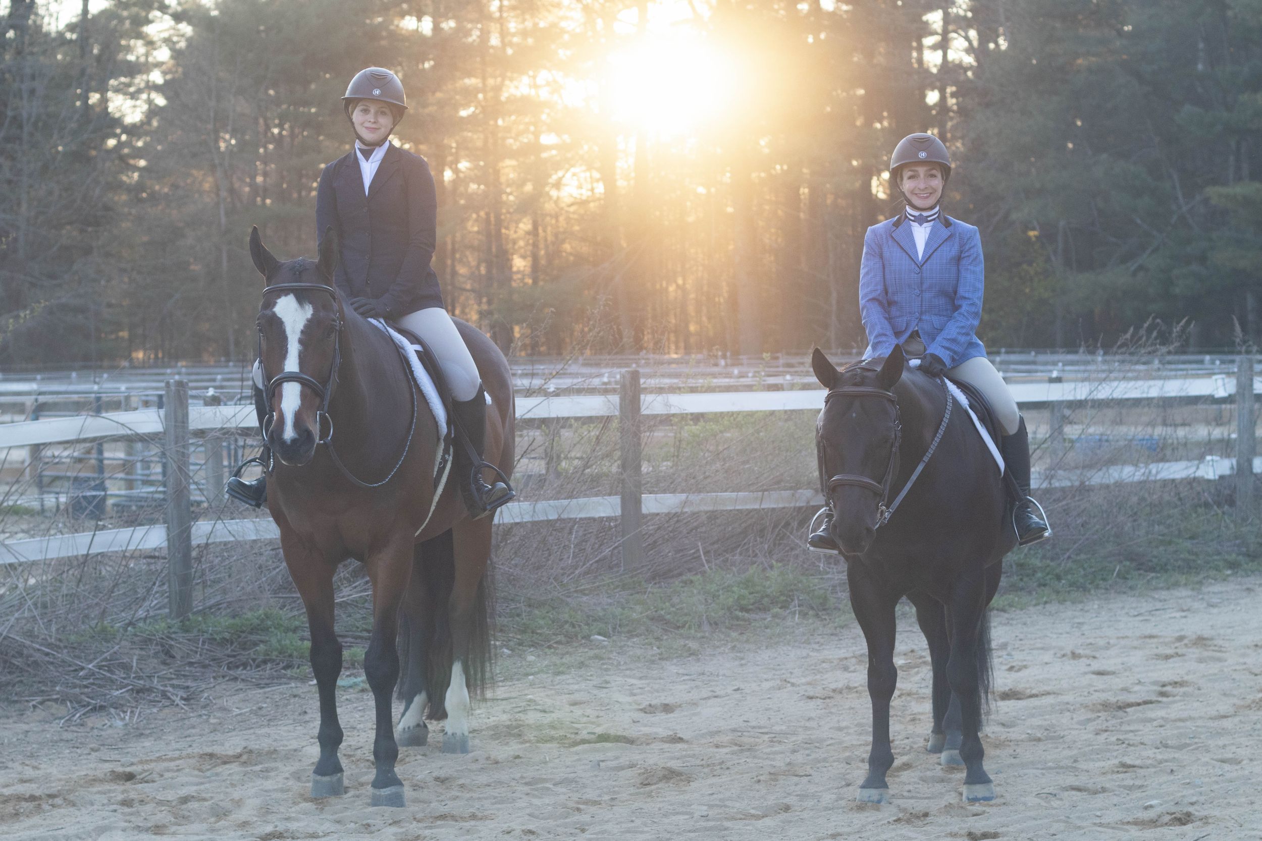 Two riders on dark horses in sandy arena at golden sunset hour.
