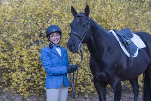 Young equestrian in blue jacket poses with black horse wearing bridle outdoors.