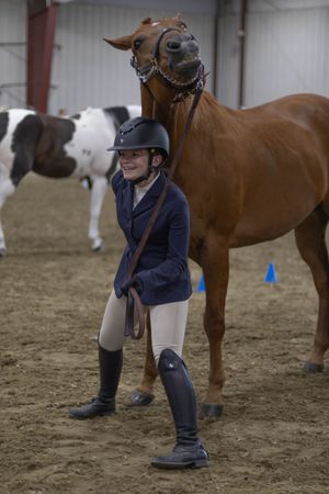 Young equestrian in navy jacket and helmet smiles beside chestnut horse in sandy indoor riding arena.
