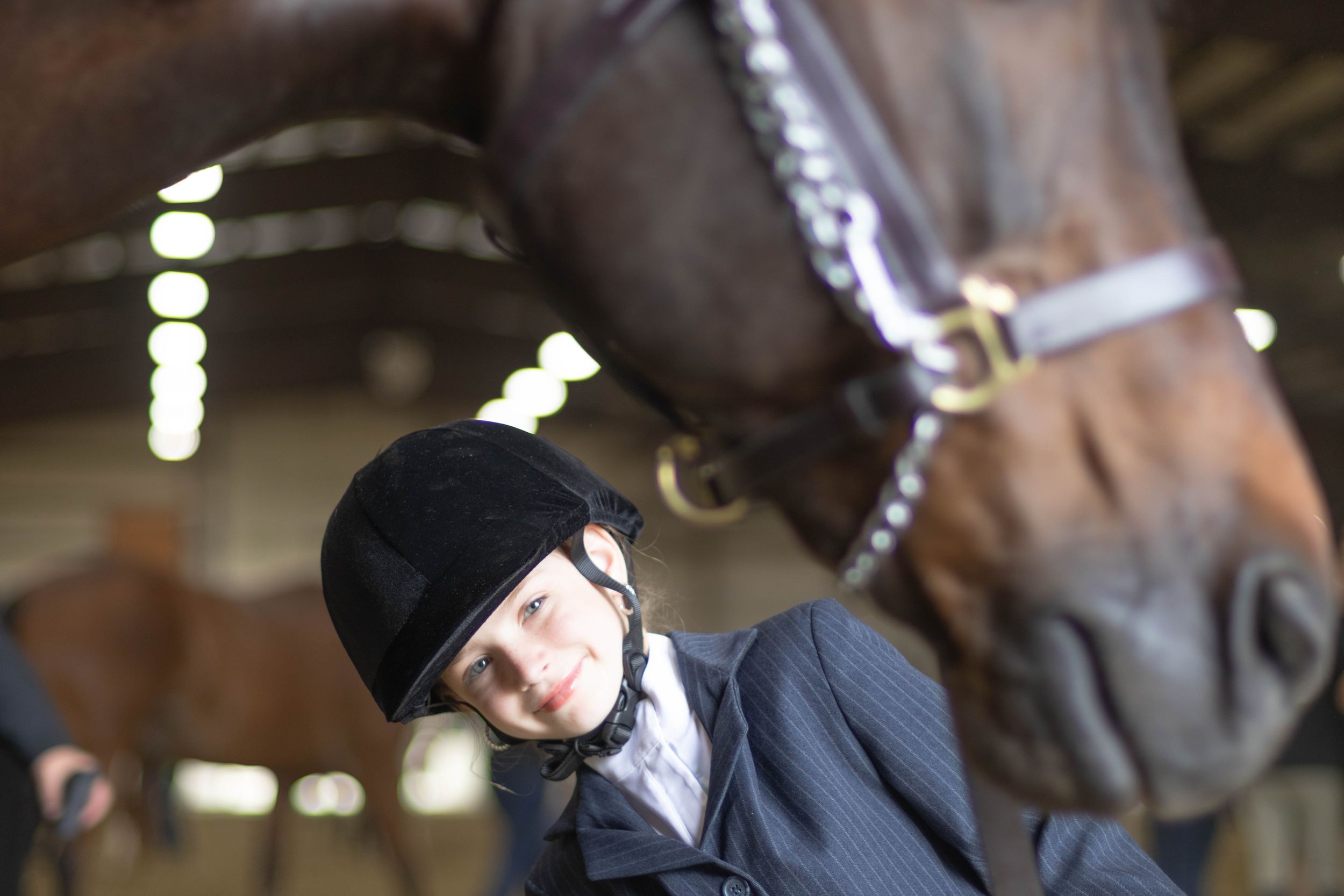 Young equestrian rider in black velvet helmet sitting beside a chestnut horse in an indoor stable.